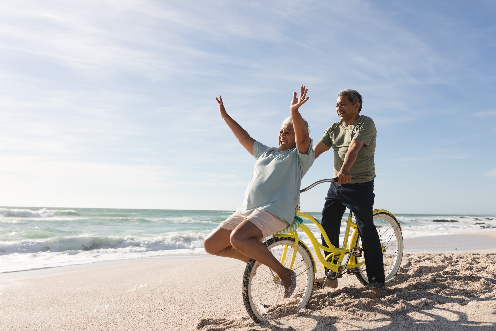 Cheerful senior multiracial couple enjoying retirement with bicycle at beach on sunny day